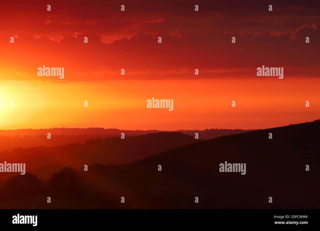 Il sole che tramonta riempie il cielo di caldo colore arancione mentre i raggi di luce solare cadono sugli strati oscuri e ondulati della campagna del Dorset. Foto Stock