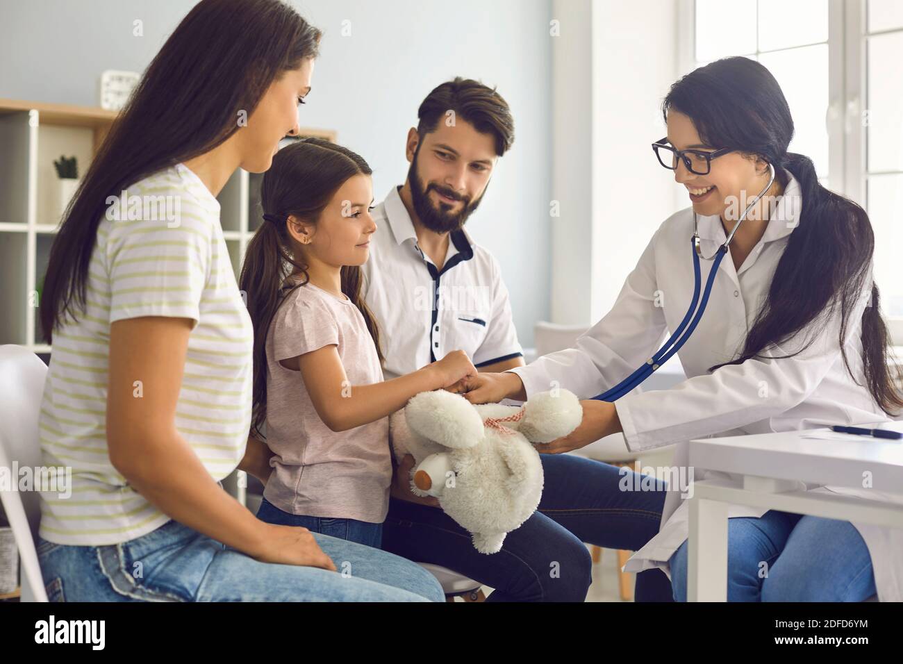 Il medico di famiglia ascolta con uno stetoscopio il bambino con i genitori seduti al tavolo della clinica. Foto Stock