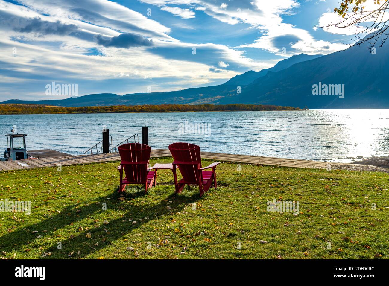 La sedia rossa guarda sopra il lancio della barca del lago di Waterton centrale nella mattina di stagione del fogliame autunnale. Luce del sole che passa il cielo blu e le nuvole su Mountains.Waterton Lake Foto Stock