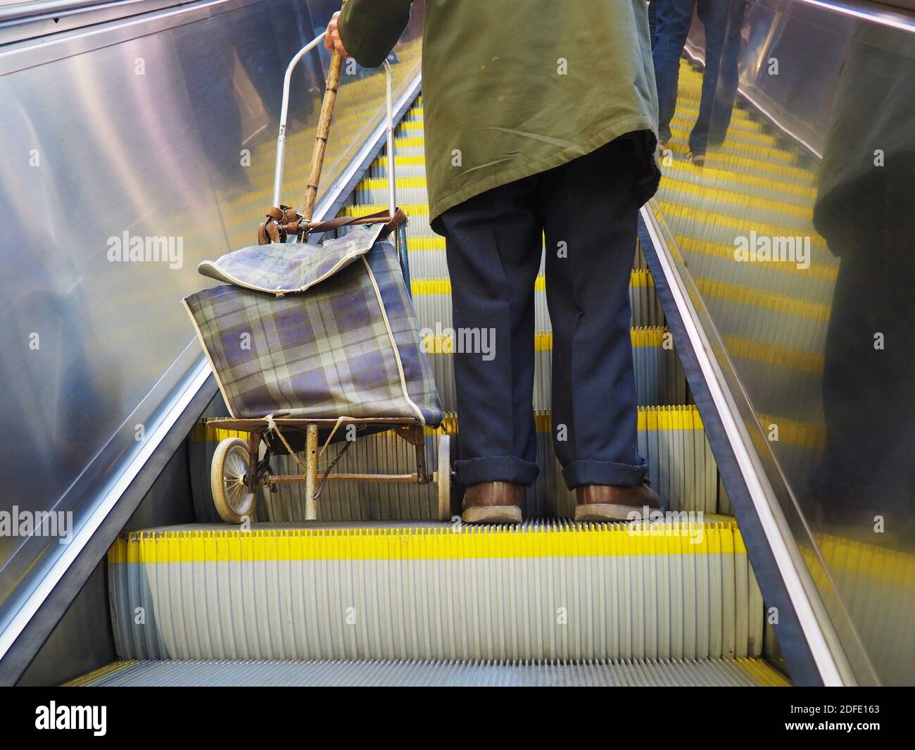 Anziano povero pensionato pensionato uomo in piedi con il carrello della spesa e bastone sulla scala mobile della metropolitana a Budapest, Ungheria Foto Stock