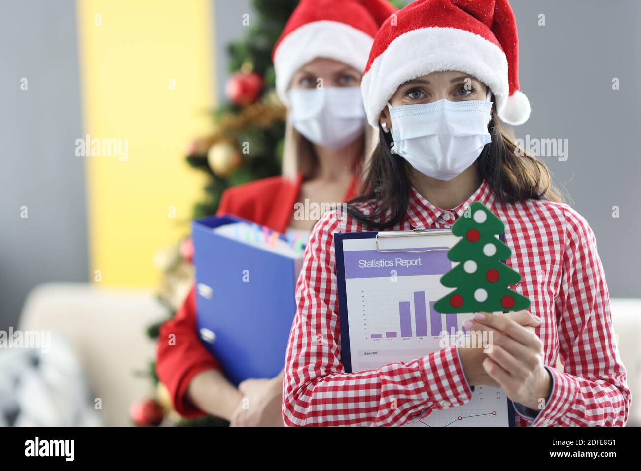 Due donne che indossano maschere di protezione medica e cappelli babbo natale cartelle con documenti e piccolo albero di natale Foto Stock