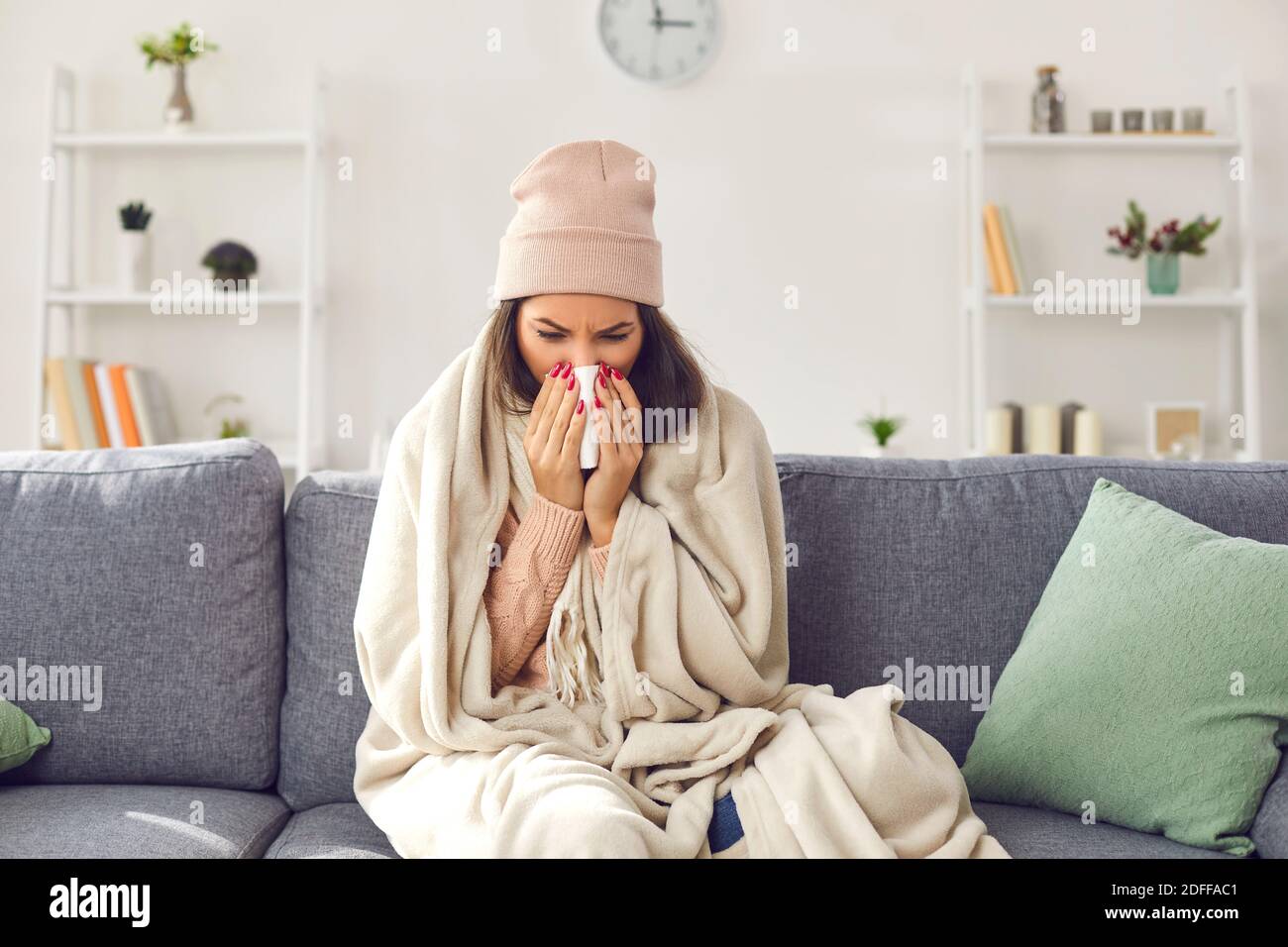 Giovane donna malata in abiti caldi, cappello seduto sul divano sotto la coperta, naso soffiato e sensazione di malessere Foto Stock