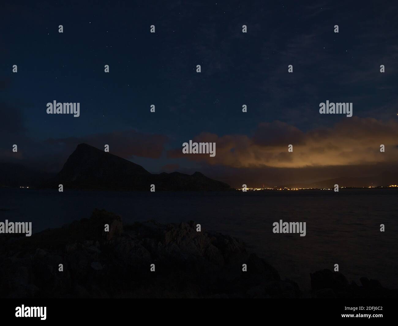 Bella vista notturna della costa occidentale dell'isola di Vestvågøy con stelle sul cielo, le sagome delle montagne e delle nuvole illuminate dalle luci della città. Foto Stock