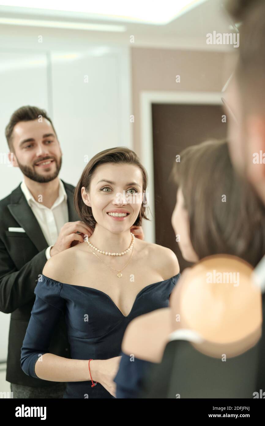 Felice giovane bella donna in abito nero sera in piedi dentro davanti allo specchio e guardando il suo marito elegante aiutare con collana Foto Stock