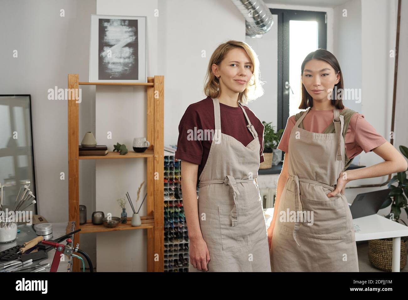 Due giovani donne di successo interculturali in abbigliamento da lavoro in piedi vicino a. uno di fronte alla telecamera in un'officina di grandi dimensioni o. studio Foto Stock
