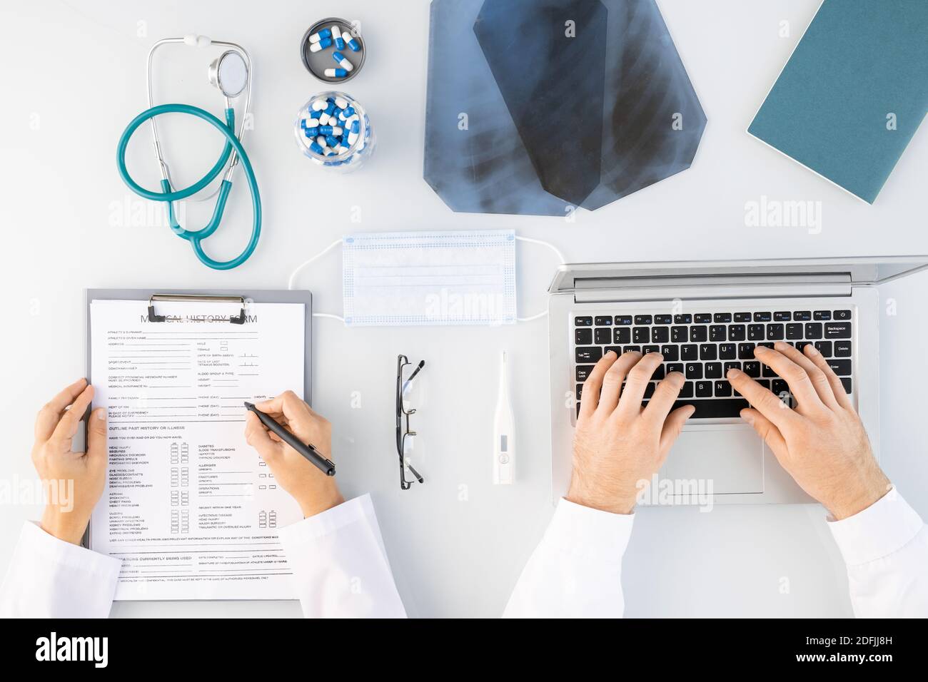 Flatlay di mani di medici di sesso maschile e femminile che fanno note mediche e la digitazione da computer portatile circondato da immagini radiografiche, pillole e stetoscopio Foto Stock