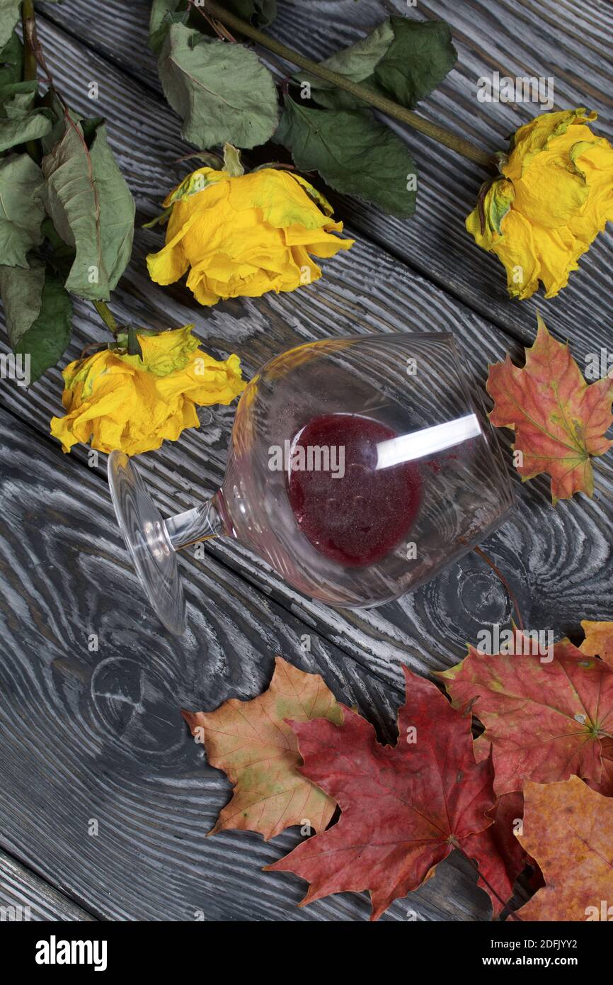 Un bouquet di rose gialle secche e foglie d'autunno secche. Un bicchiere rovesciato di vino affolto. Su tavole di pino. Foto Stock