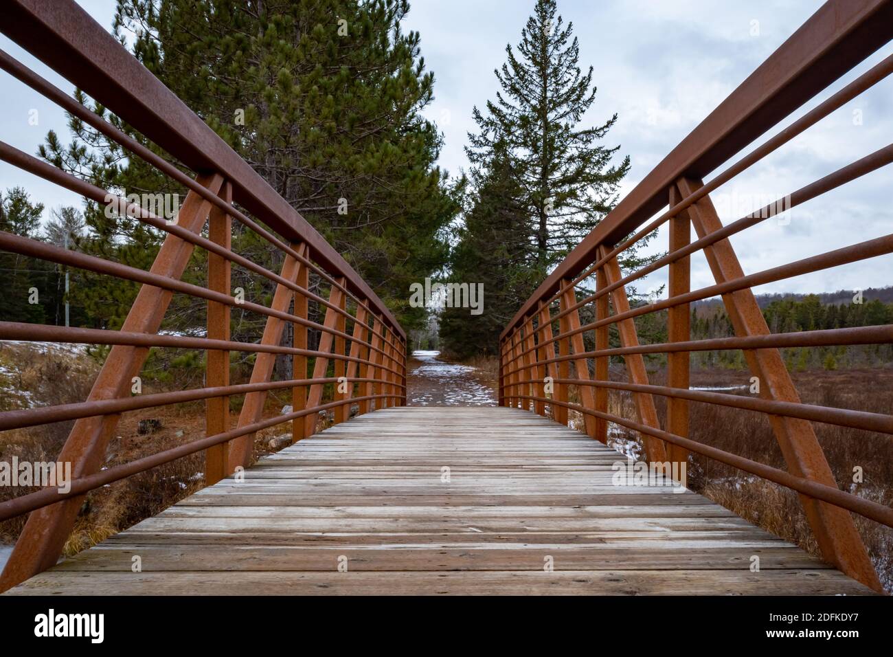 Una vista dal centro di un ponte pedonale in legno su un piccolo ruscello. L'acciaio resistente agli agenti atmosferici forma ringhiere che si estendono su entrambi i lati. Neve leggera, ev Foto Stock