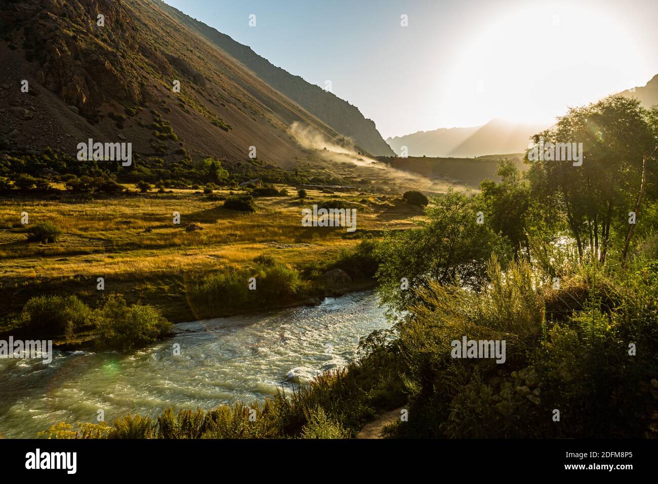 Un fiume scorre attraverso la valle vicino a Sarytag, Tagikistan, mentre la polvere si alza sulla strada di accesso in lontananza e il sole basso proietta lunghe ombre. Foto Stock