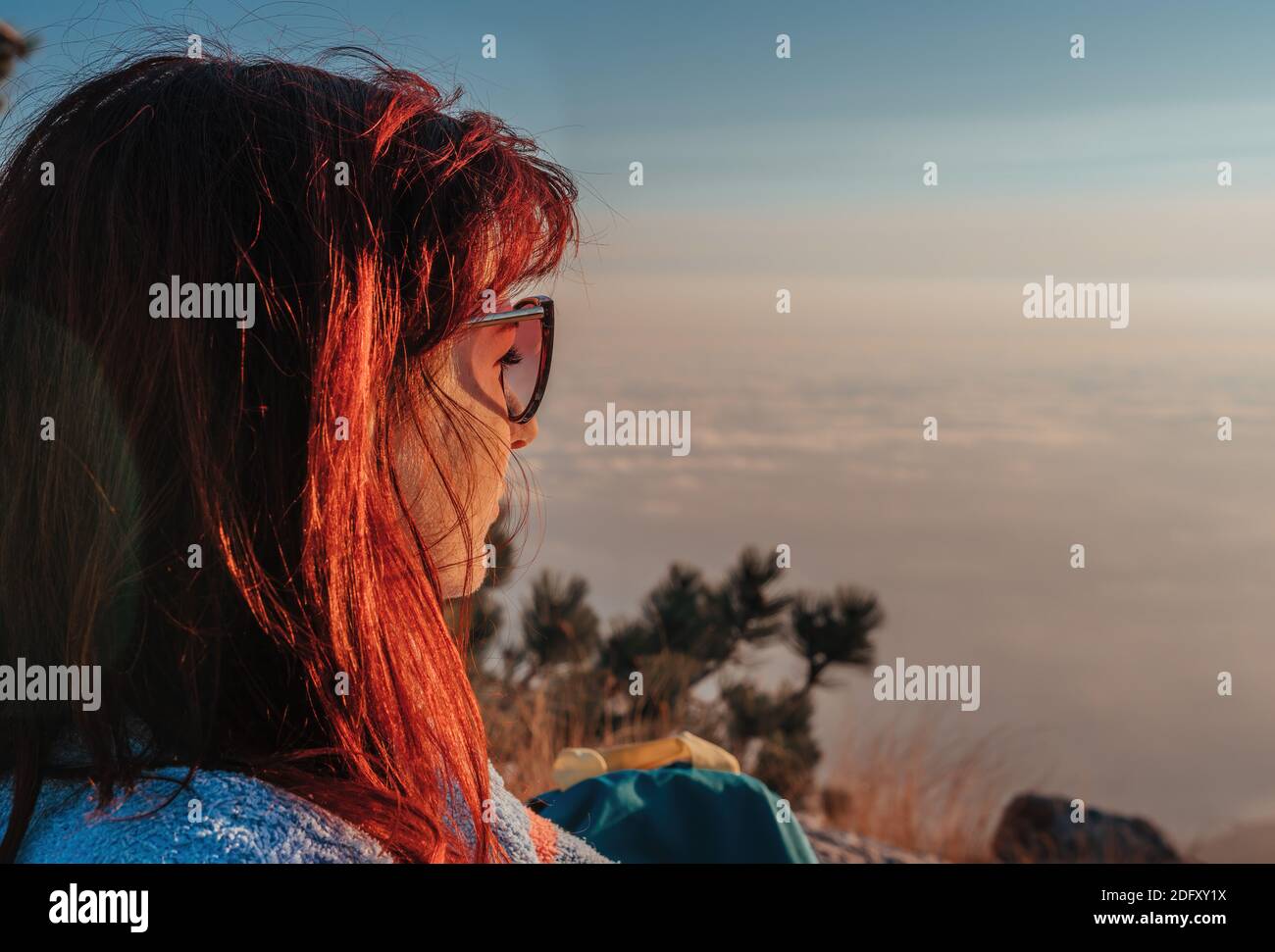 Giovane donna rossa in sassolini seduta su una roccia in cima alle montagne, sopra le nuvole e godendosi il caldo tramonto. Spazio di copia. Il concetto di Foto Stock