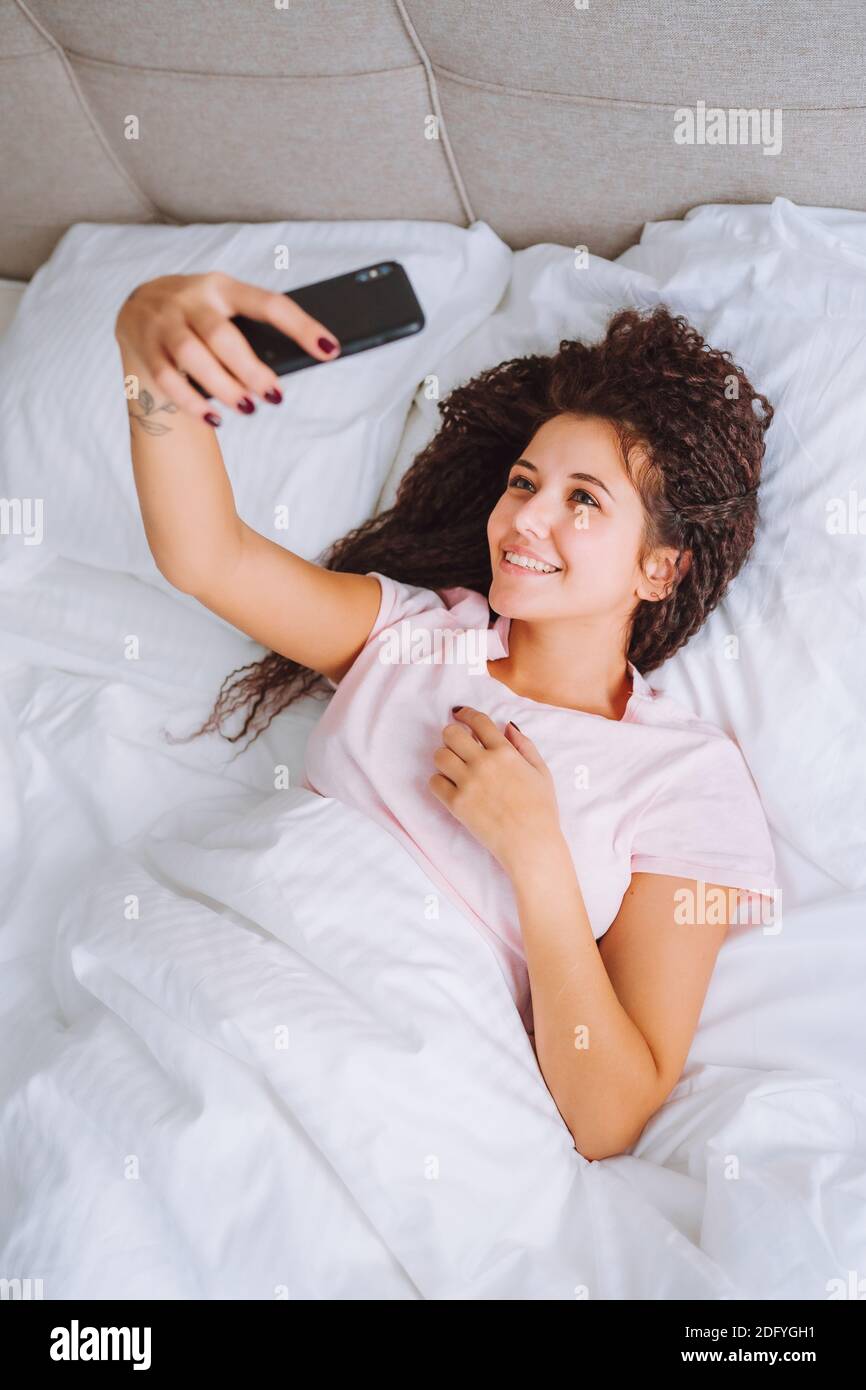 Giovane donna sorridente afro capelli sdraiati nel letto e utilizzare smartphone facendo selfie. Dipendenza dai social media, datazione online. Foto Stock