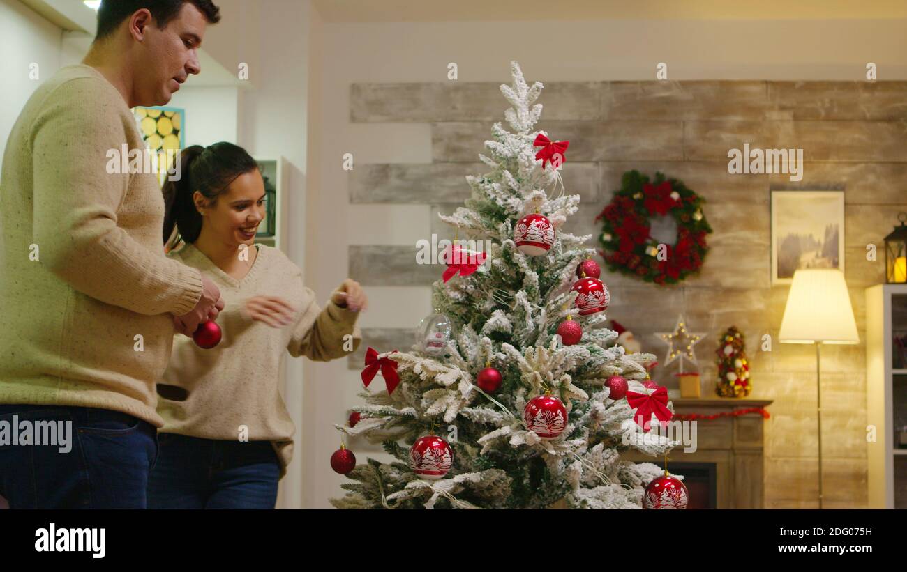 La coppia con i vestiti corrispondenti decorano il loro albero di natale per le feste invernali. Decorazione bello albero di Natale con decorazioni palla di vetro. Moglie e marito in abiti coordinati che aiutano a casa ornata con luci di ghirlanda Foto Stock