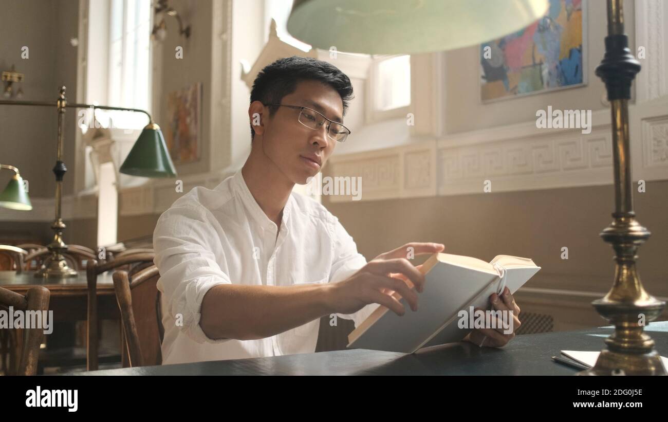 Giovane studente asiatico maschile che studia con attenzione con il libro in biblioteca universitaria. Ragazzo sicuro che si prepara per gli esami all'università Foto Stock