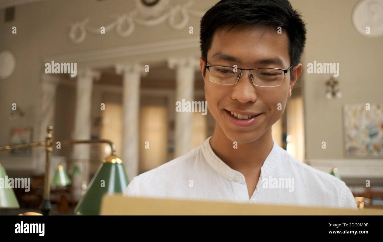 Ritratto di sorridente studente asiatico felice apertura busta con risultati esame in biblioteca universitaria. Giovane ragazzo attraente che studia all'università Foto Stock