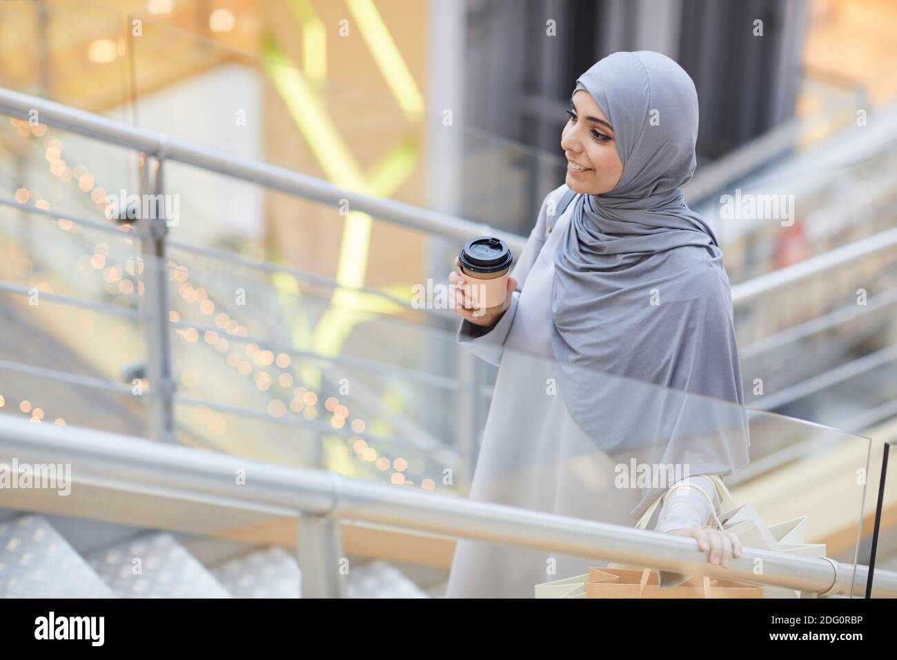 Ritratto di sorridente donna medio-orientale che tiene la tazza di caffè mentre salendo le scale nel centro commerciale, copy space Foto Stock