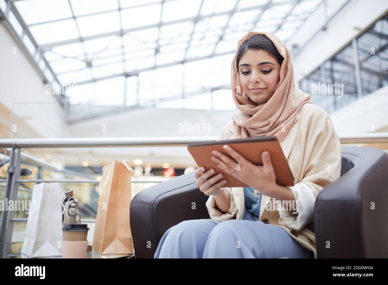 Ritratto della moderna donna mediorientale che indossa un copricapo e utilizza un tablet digitale mentre si siede nel centro commerciale, copia spazio Foto Stock