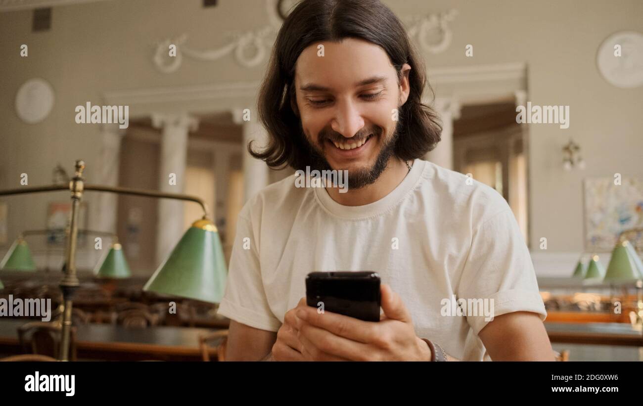 Ritratto di allegro studente casual felicemente utilizzando smartphone in libreria. Ragazzo attraente con telefono che studia all'università Foto Stock