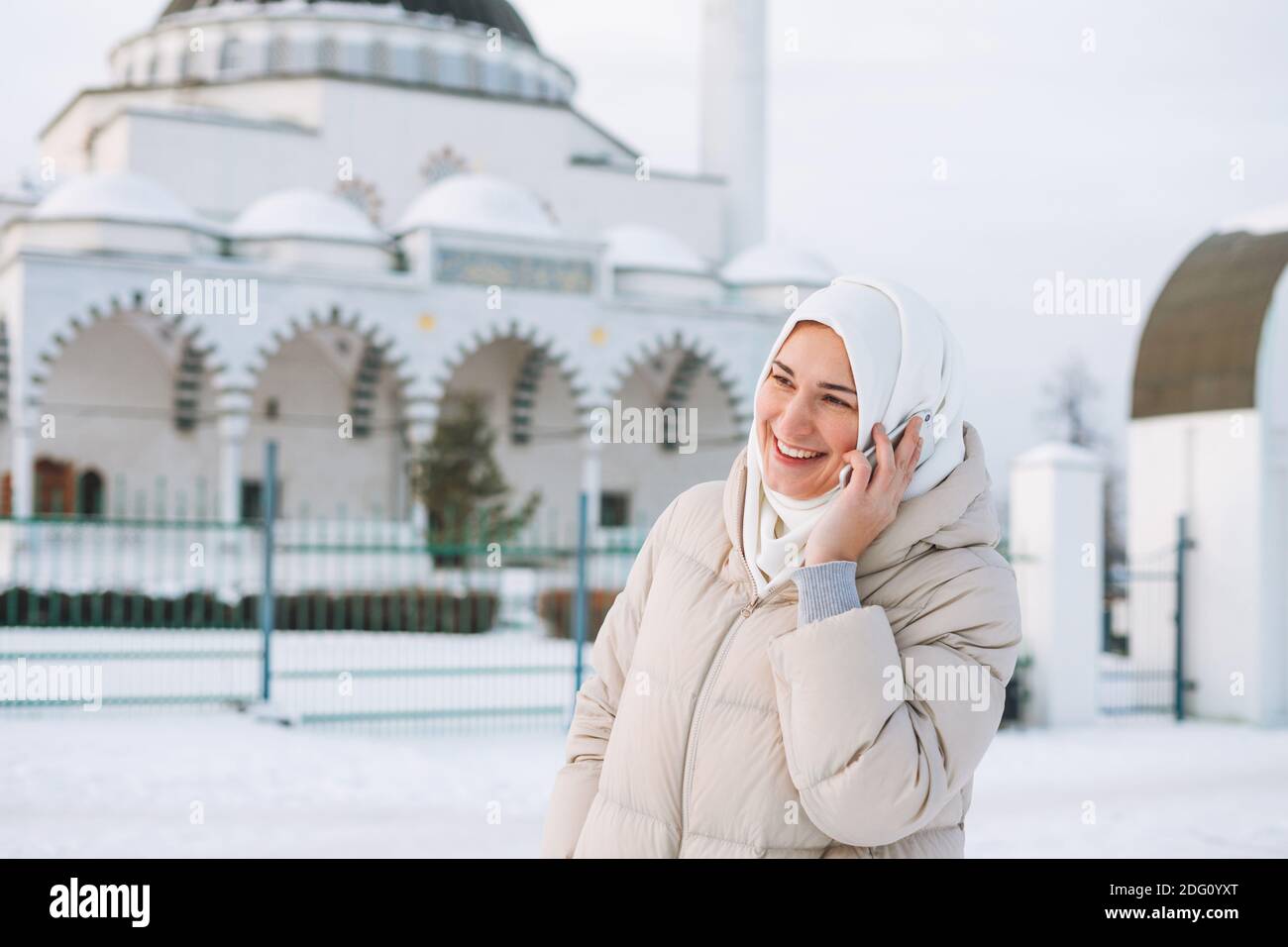 Bella giovane donna musulmana sorridente in foulard in abiti leggeri utilizzo di dispositivi mobili sullo sfondo della moschea nella stagione invernale Foto Stock