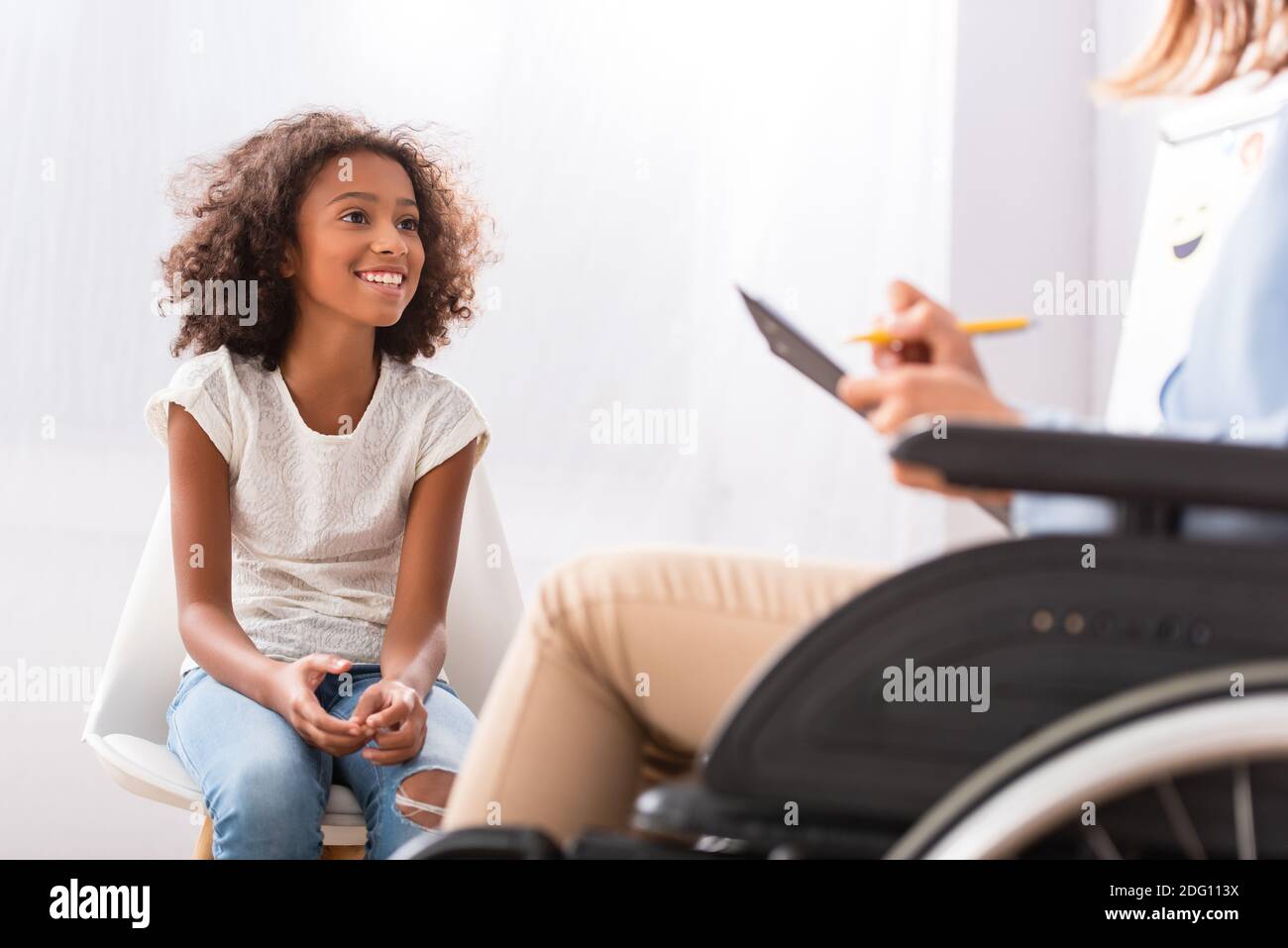 Felice afroamericana ragazza seduta su sedia con psicologo offuscato in sedia a rotelle in primo piano Foto Stock