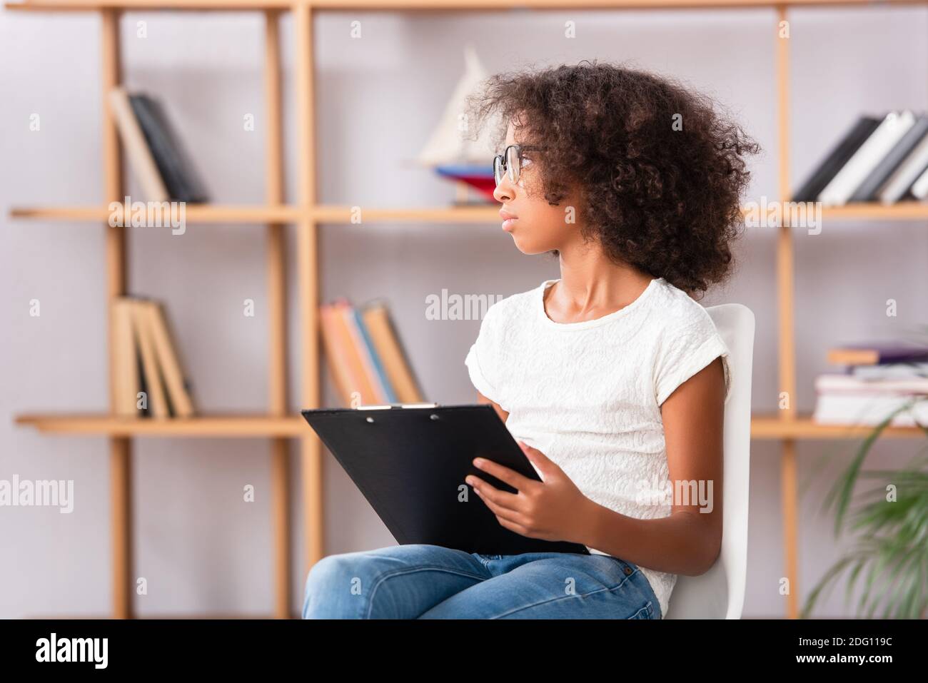 ragazza afroamericana in occhiali con appunti guardando via mentre seduta sulla sedia su sfondo sfocato Foto Stock