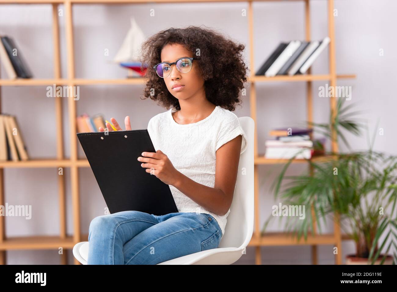 Ragazza afroamericana in occhiali con appunti guardando via mentre seduta sulla sedia su sfondo sfocato Foto Stock