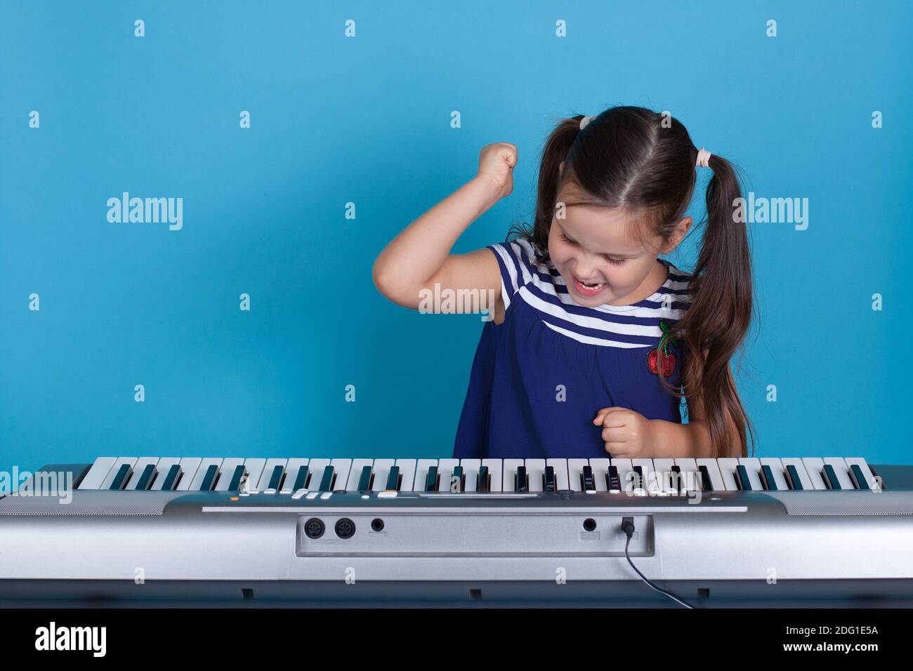 primo piano bambina in un vestito blu batte i suoi pugni sui tasti del sintetizzatore con rabbia e non vuole imparare, isolato su uno sfondo blu Foto Stock