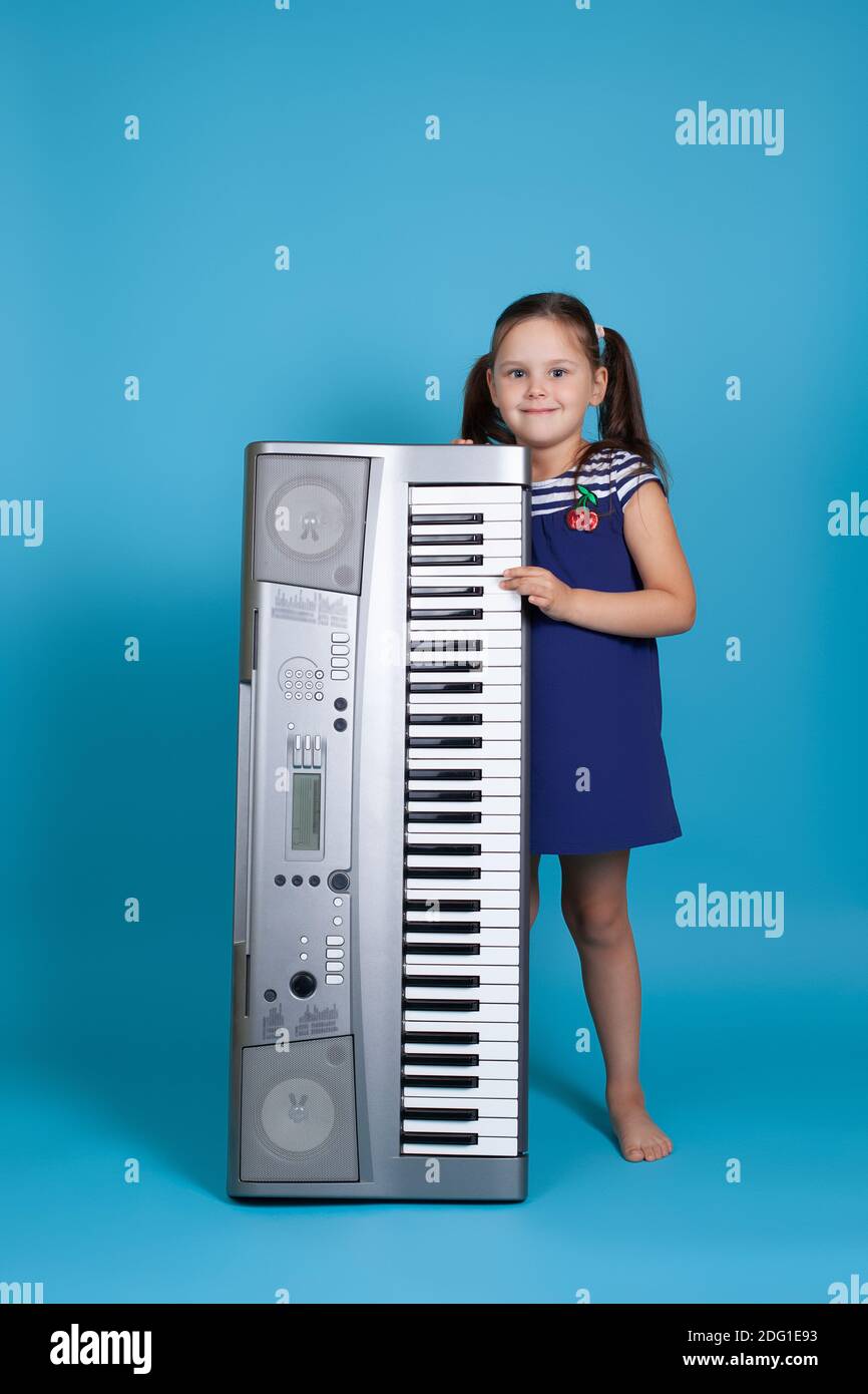 una bambina di lunghezza intera in un vestito blu tiene un sintetizzatore elettronico verticale isolato su sfondo blu Foto Stock
