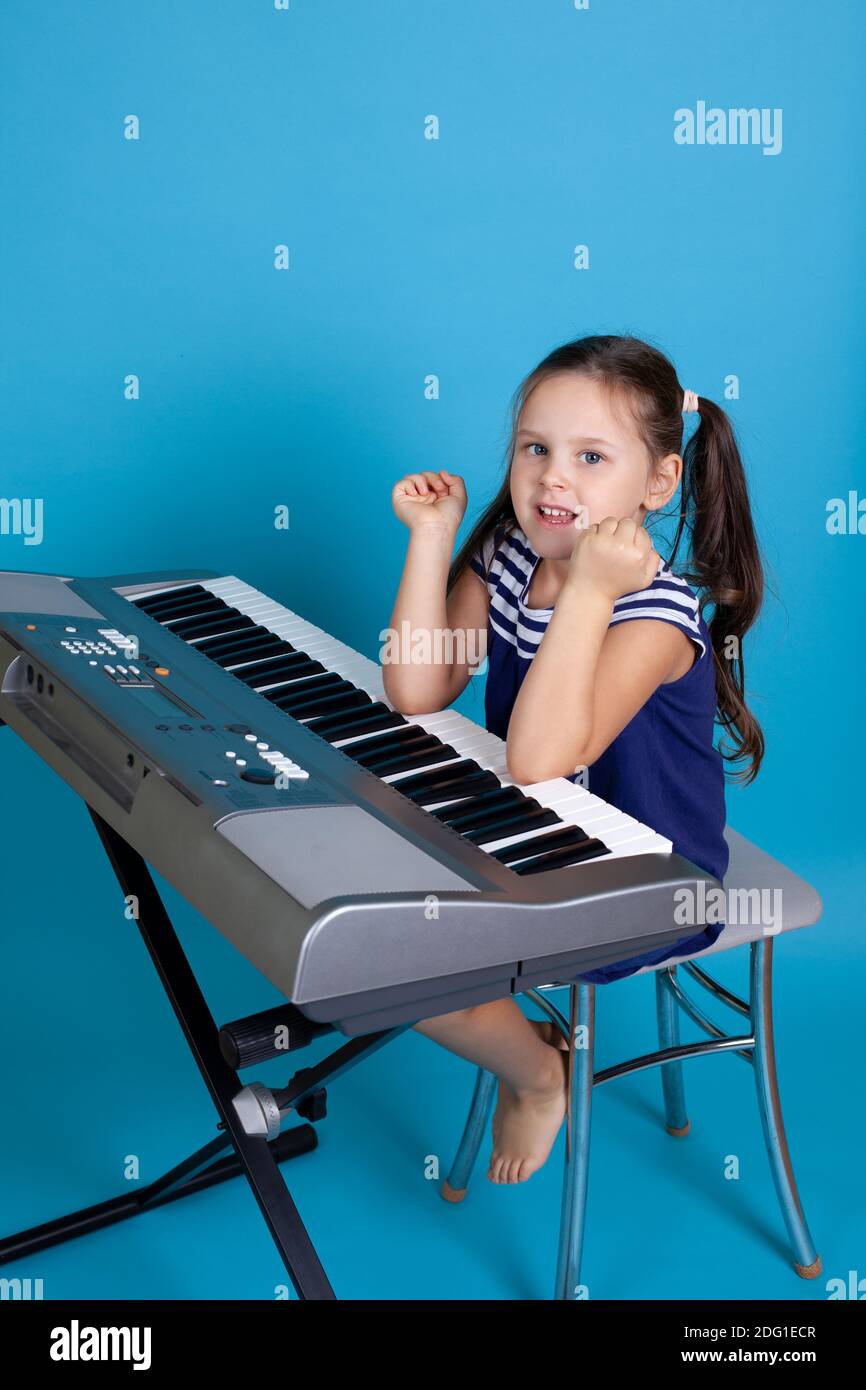 una bambina a lunghezza intera in un abito blu si siede su una sedia e suona il pianoforte con i suoi gomiti, caratteristiche alternative isolate su uno sfondo blu Foto Stock