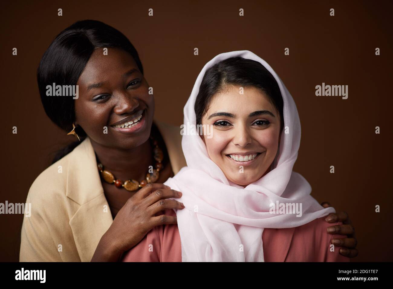 Ritratto minimo di due giovani donne etniche che sorridono alla macchina fotografica mentre si posa contro sfondo marrone in studio e celebrare il patrimonio e ide personale Foto Stock