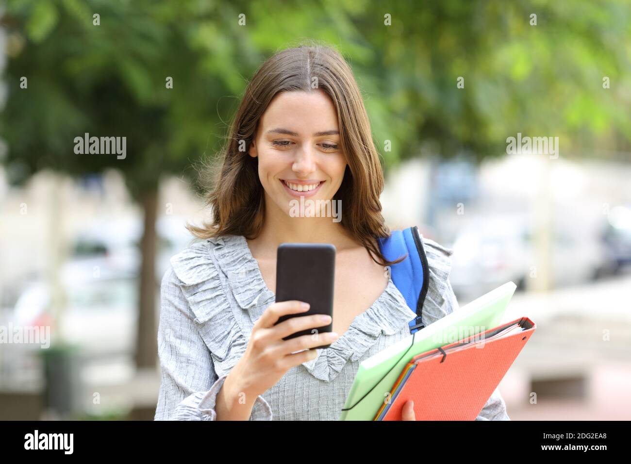 Vista frontale ritratto di uno studente felice che cammina controllando intelligente telefono in strada Foto Stock
