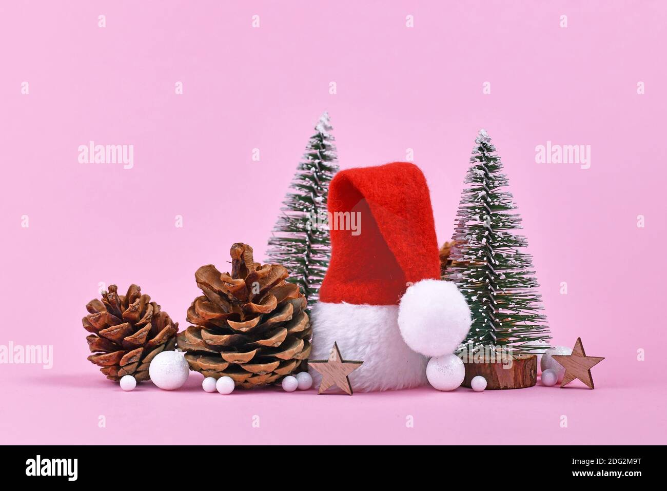 Arrangiamento di Natale con cappello rosso di Santa, cono di pino, ornamenti stellari e piccoli alberi di Natale su sfondo rosa Foto Stock