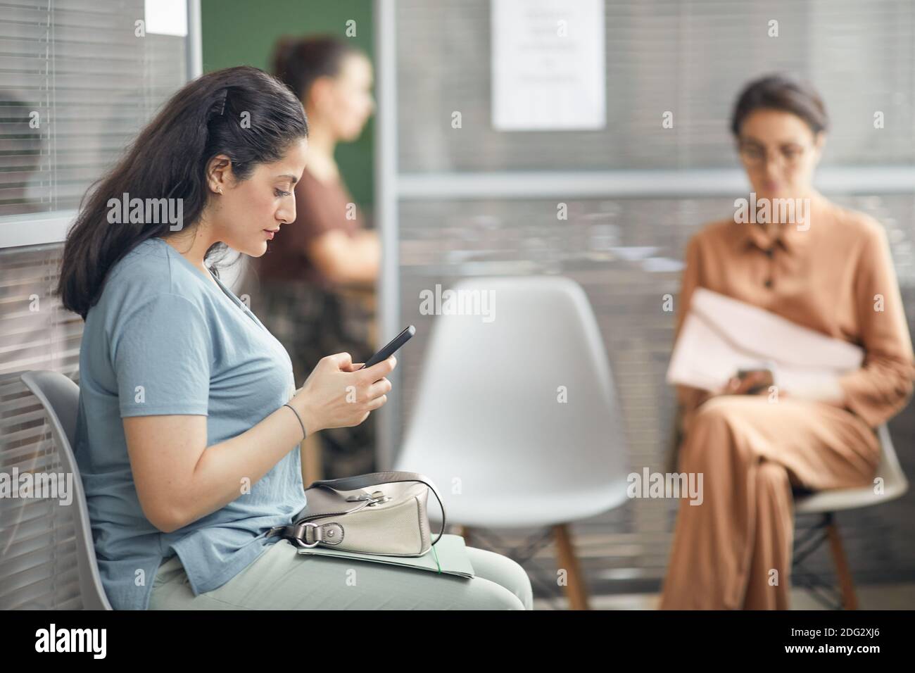 Vista laterale ritratto di giovane donna nervosa guardando smartphone mentre in attesa in coda per il colloquio di lavoro in ufficio, spazio di copia Foto Stock