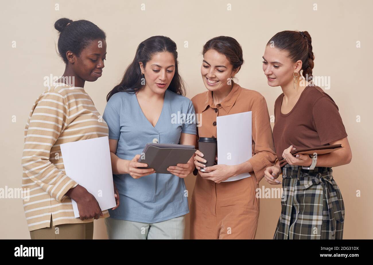 Gruppo multietnico di quattro giovani donne d'affari che guardano al tablet digitale mentre si è in piedi a muro in ufficio Foto Stock