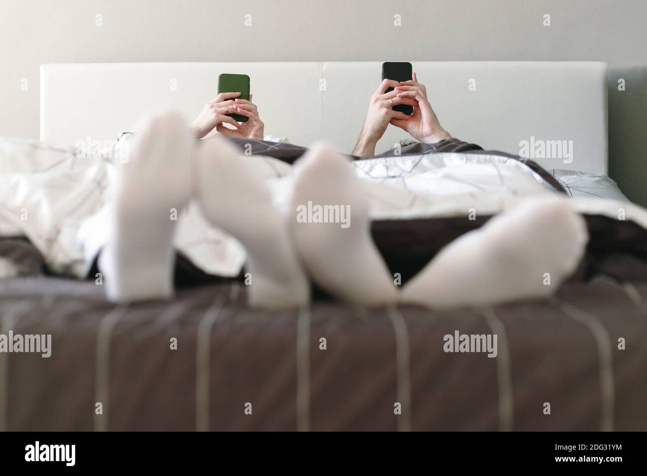 La giovane famiglia a letto al mattino si trova con gli smartphone. Persone sposate con dipendenza da Internet dai social network. Foto Stock