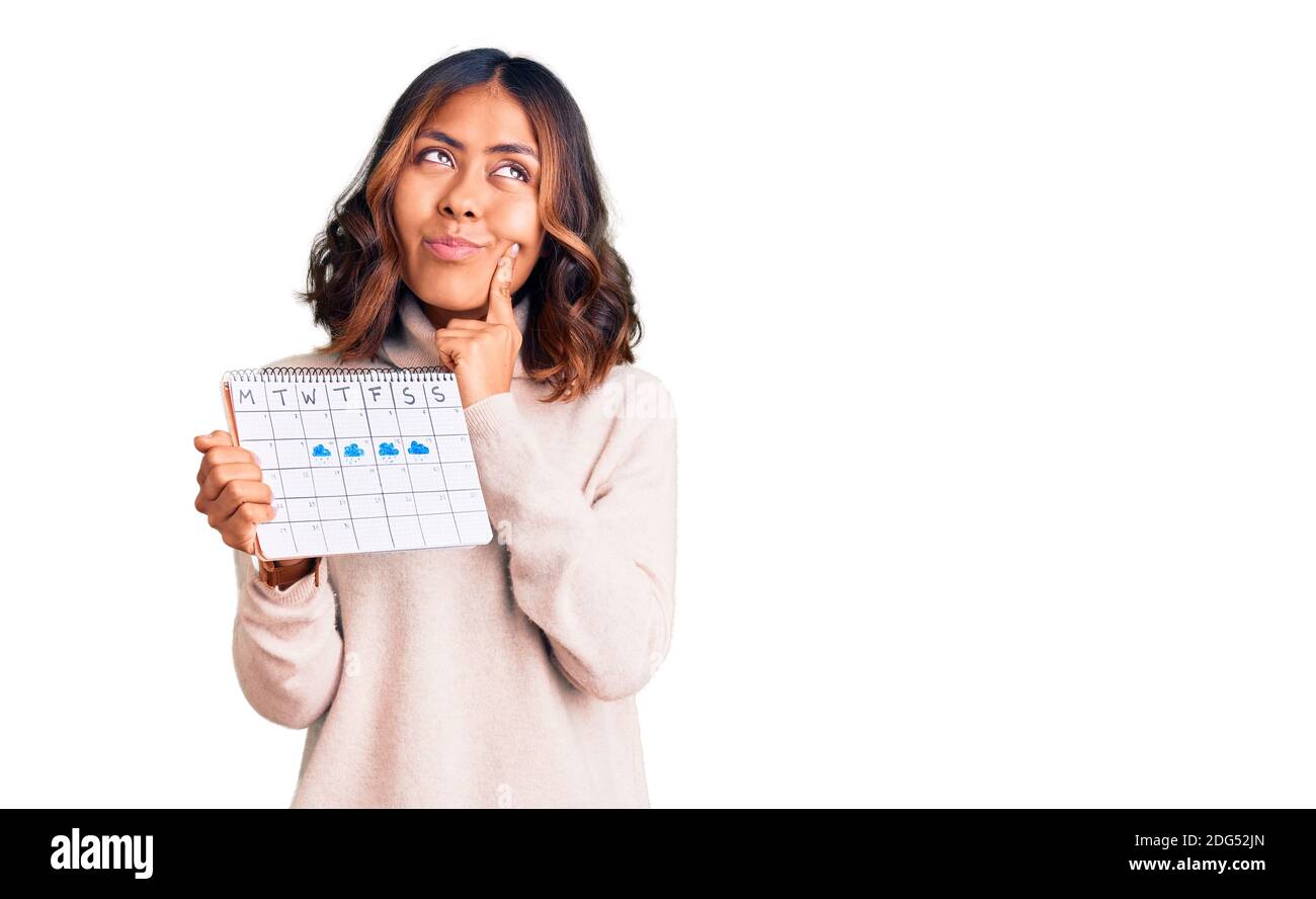Giovane bella razza mista donna tenendo calendario di viaggio faccia seria pensare alla domanda con la mano sul mento, pensieroso circa idea confusa Foto Stock