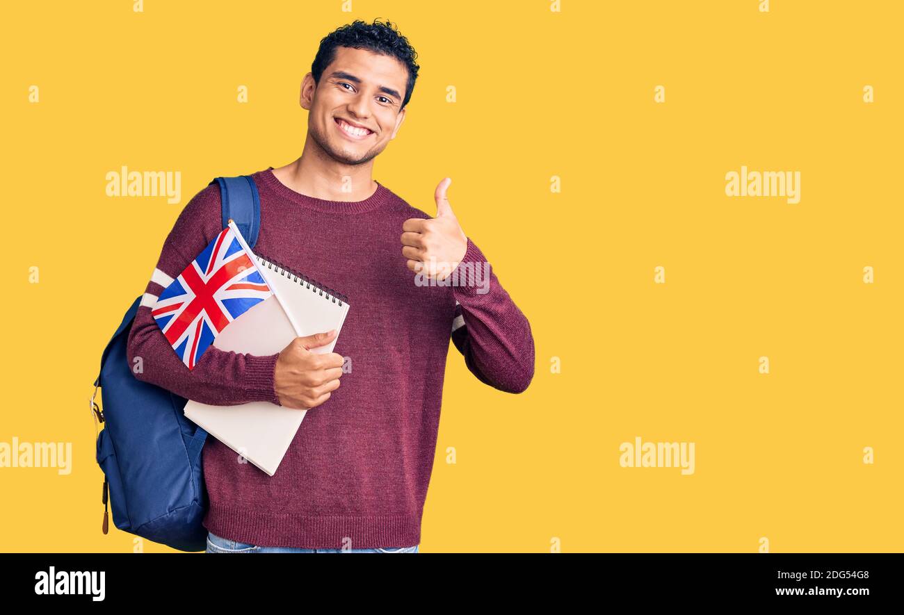 Hispanic bello giovane uomo scambio studente tenendo bandiera uk sorridente felice e positivo, pollice su facendo eccellente e segno di approvazione Foto Stock