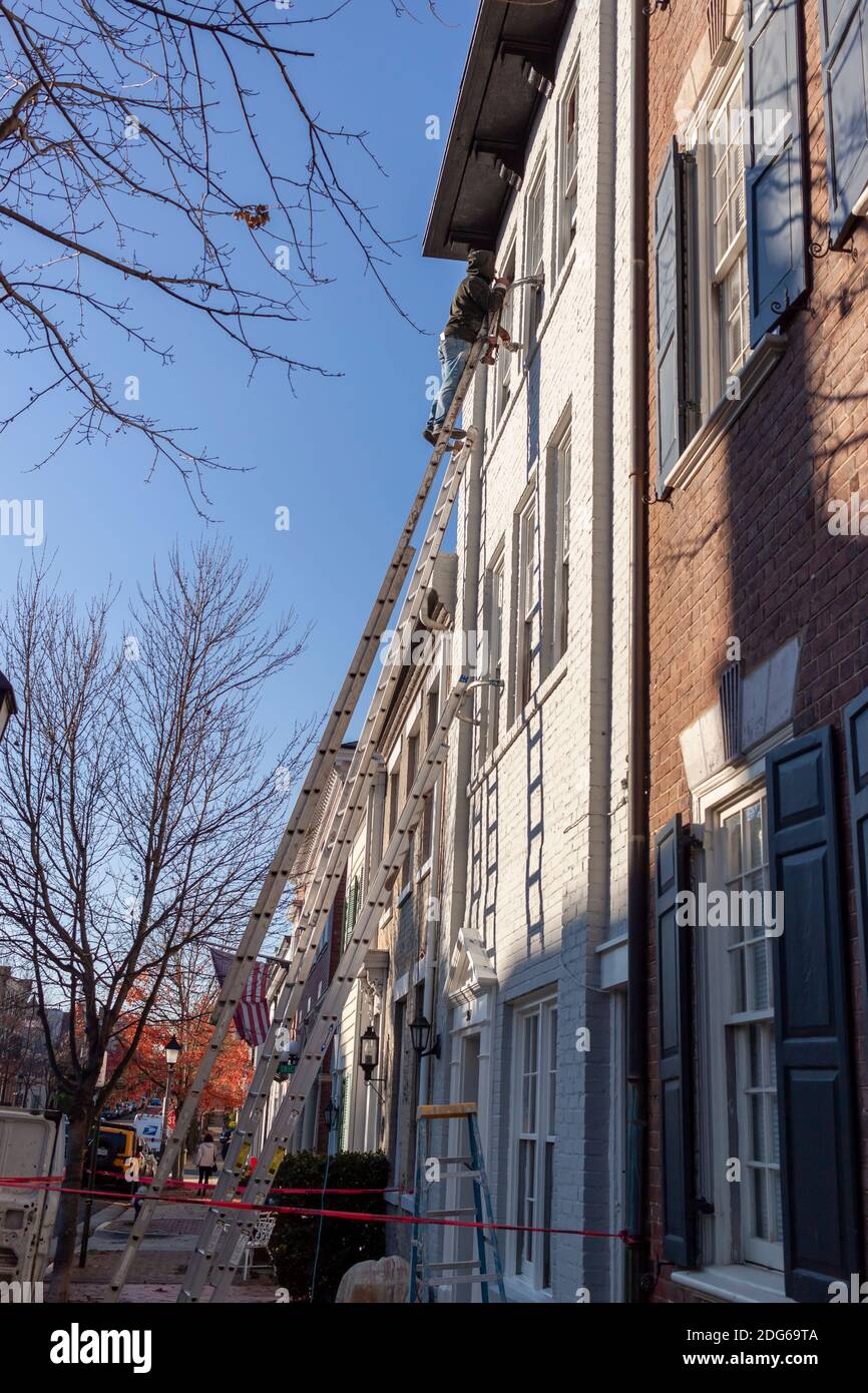 Alexandria, VA, USA 11-28-2020: Un pittore di casa è sulla parte superiore di una scala di estensione di alluminio di dimensione industriale che dipinge la superficie esterna di un vecchio bri Foto Stock