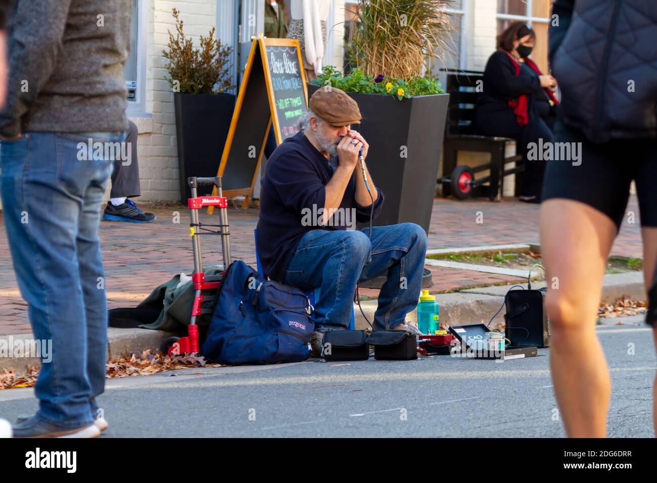 Alexandria, VA, USA 11-28-2020: Un musicista di strada anziano con cuscinetti bianchi che indossa scarpe, jeans e cappello piatto seduto sullo sgabello accanto al marciapiede sta giocando Foto Stock