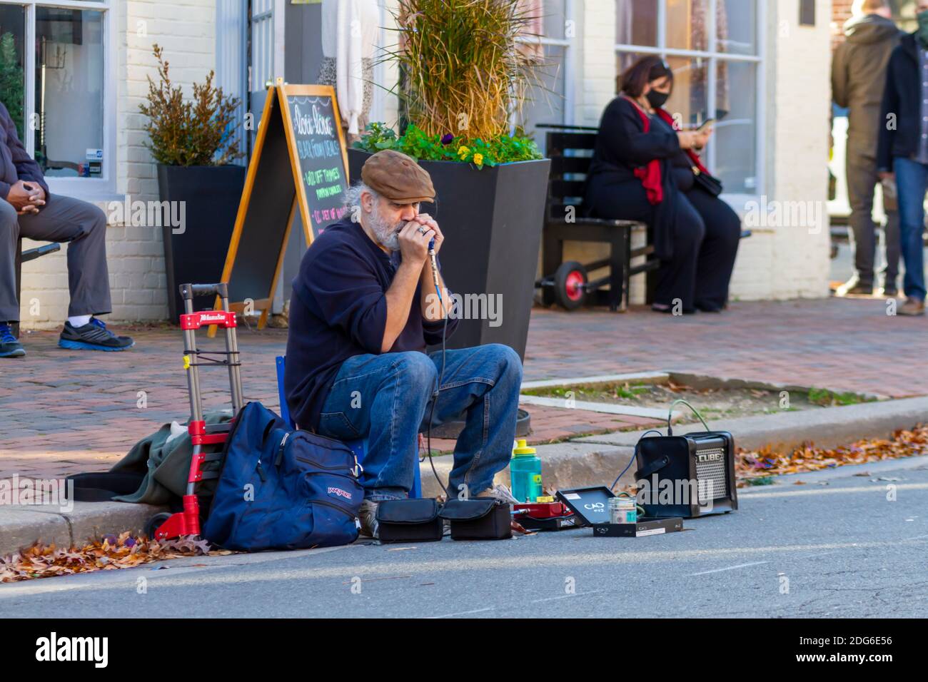 Alexandria, VA, USA 11-28-2020: Un musicista di strada anziano con cuscinetti bianchi che indossa scarpe, jeans e cappello piatto seduto sullo sgabello accanto al marciapiede sta giocando Foto Stock