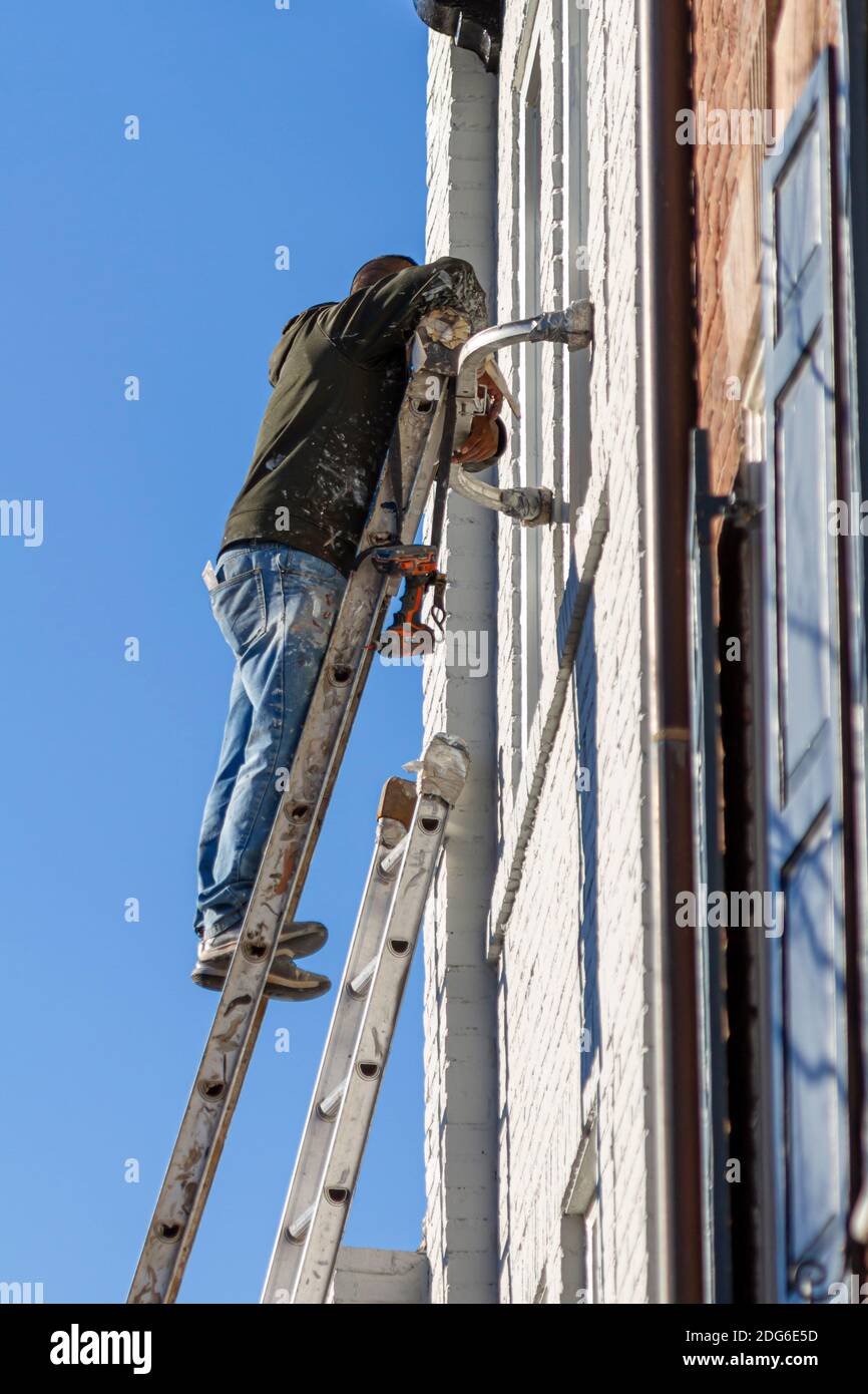 Un pittore di casa ispanica con i vestiti macchiati di vernice è sulla parte superiore di una scala di costruzione di acciaio che dipinge la superficie esterna di una vecchia casa di mattoni. Foto Stock