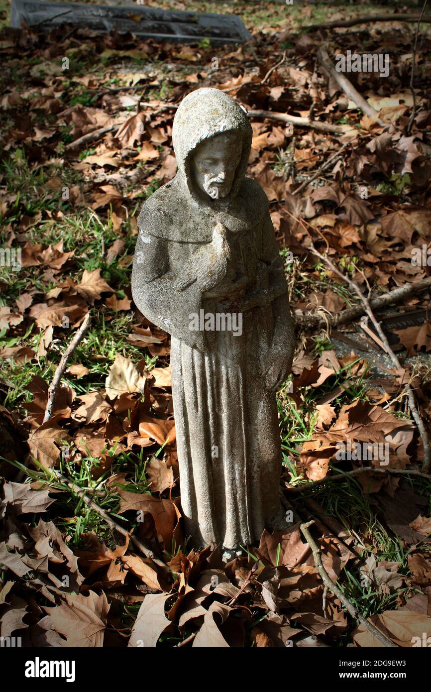 Questa piccola figurina cementizia di San Francesco d'Assisi tiene delicatamente un uccello tra le foglie secche sul terreno Foto Stock