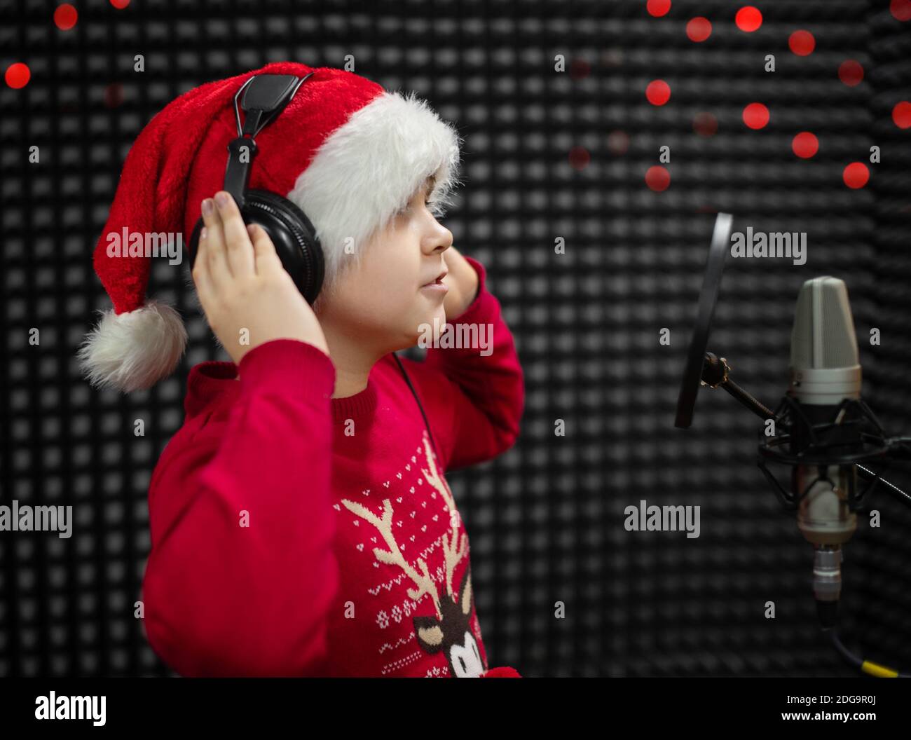 Il ragazzo in cappello di Babbo Natale e le cuffie cantano o parlano nel microfono con il filtro pop nello studio di registrazione vocale. Foto Stock