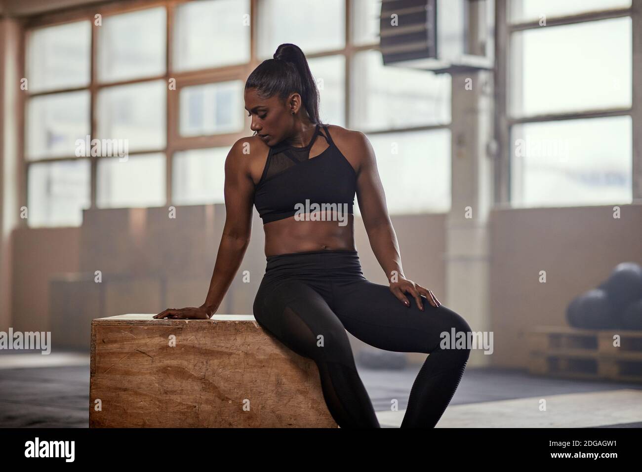 Atleta femminile di etnia muscolare in abbigliamento sportivo seduto su scatola di legno e appoggiandosi indietro mentre riposano dopo gli esercizi difficili in spazioso palestra Foto Stock