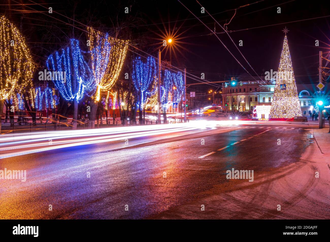 Città notturna con decorazioni natalizie, abeti e tracce di fari di automobili in movimento, riflessi nell'asfalto bagnato. Foto Stock
