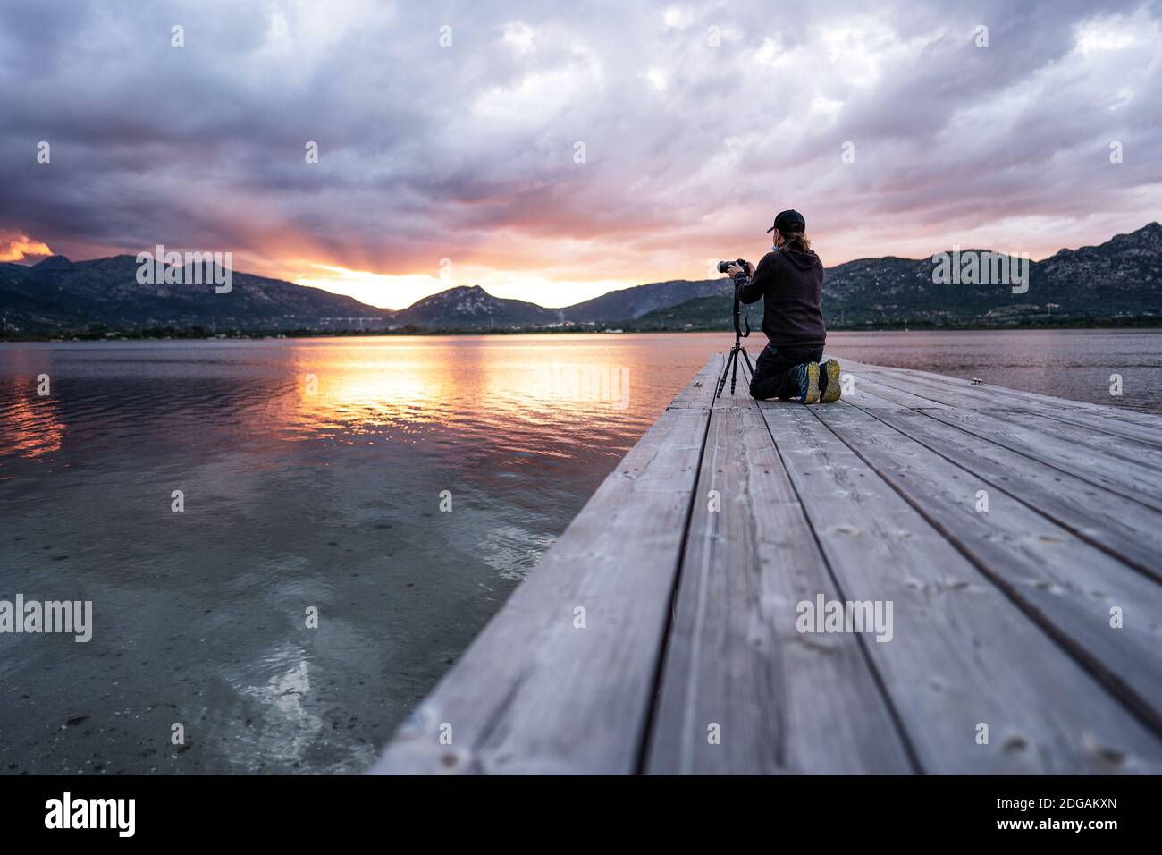 Sperimentate la natura con l'hobby della fotografia all'aperto - A. l'uomo inginocchiato su un molo di legno di un lago si regola la sua fotocamera mira a un sole nuvoloso Foto Stock