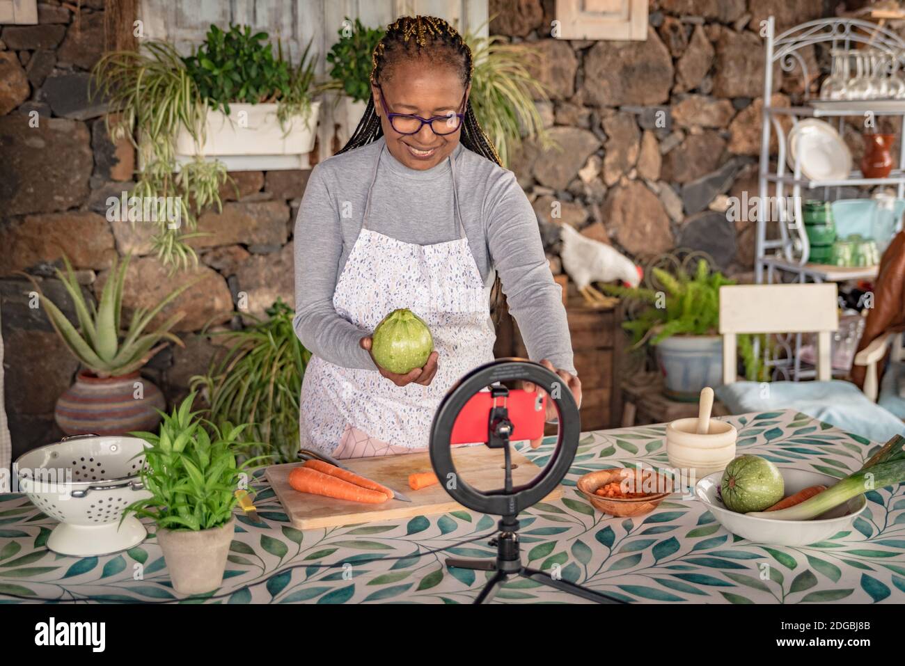 donna nera con gli occhiali insegna online con il suo telefono cellulare lezioni di cucina da casa Foto Stock