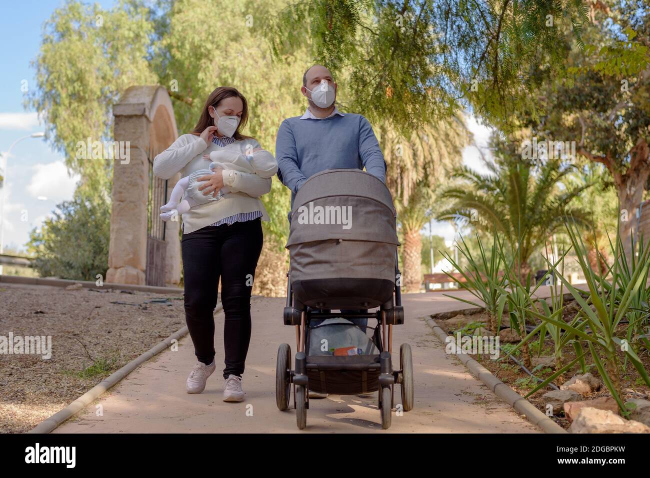 Felice coppia a piedi nel parco con passeggino. Indossano maschere mediche per proteggerle dal virus. Foto Stock