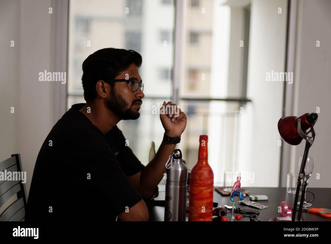 Un giovane indiano bengalese alto, scuro, bella bruna per la barba in una t-shirt casual è seduto su un tavolo da ufficio in un umore creativo. Foto Stock