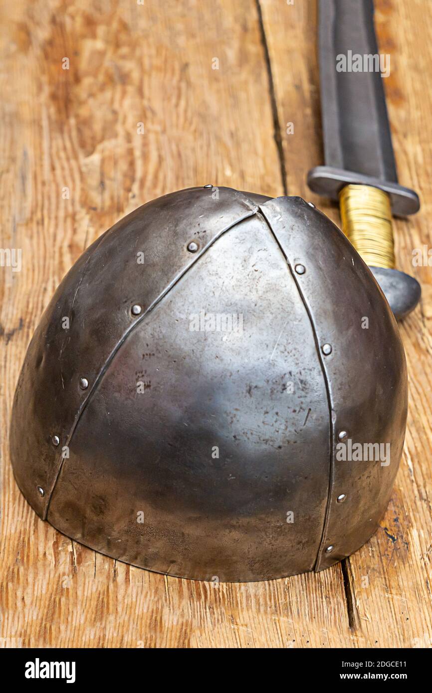 Casco in acciaio di ferro si erge sullo sfondo di un lungo spada su un tavolo di legno Foto Stock