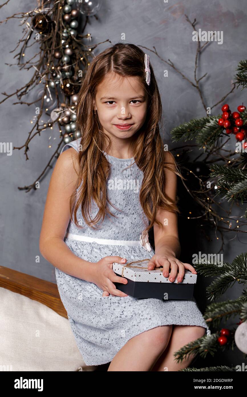 Bambina seduta vicino all'albero di Natale la vigilia di Natale, tenendo in mano la confezione regalo, guardando la macchina fotografica con espressione pensiva. Concetto di Natale o Capodanno. Foto Stock